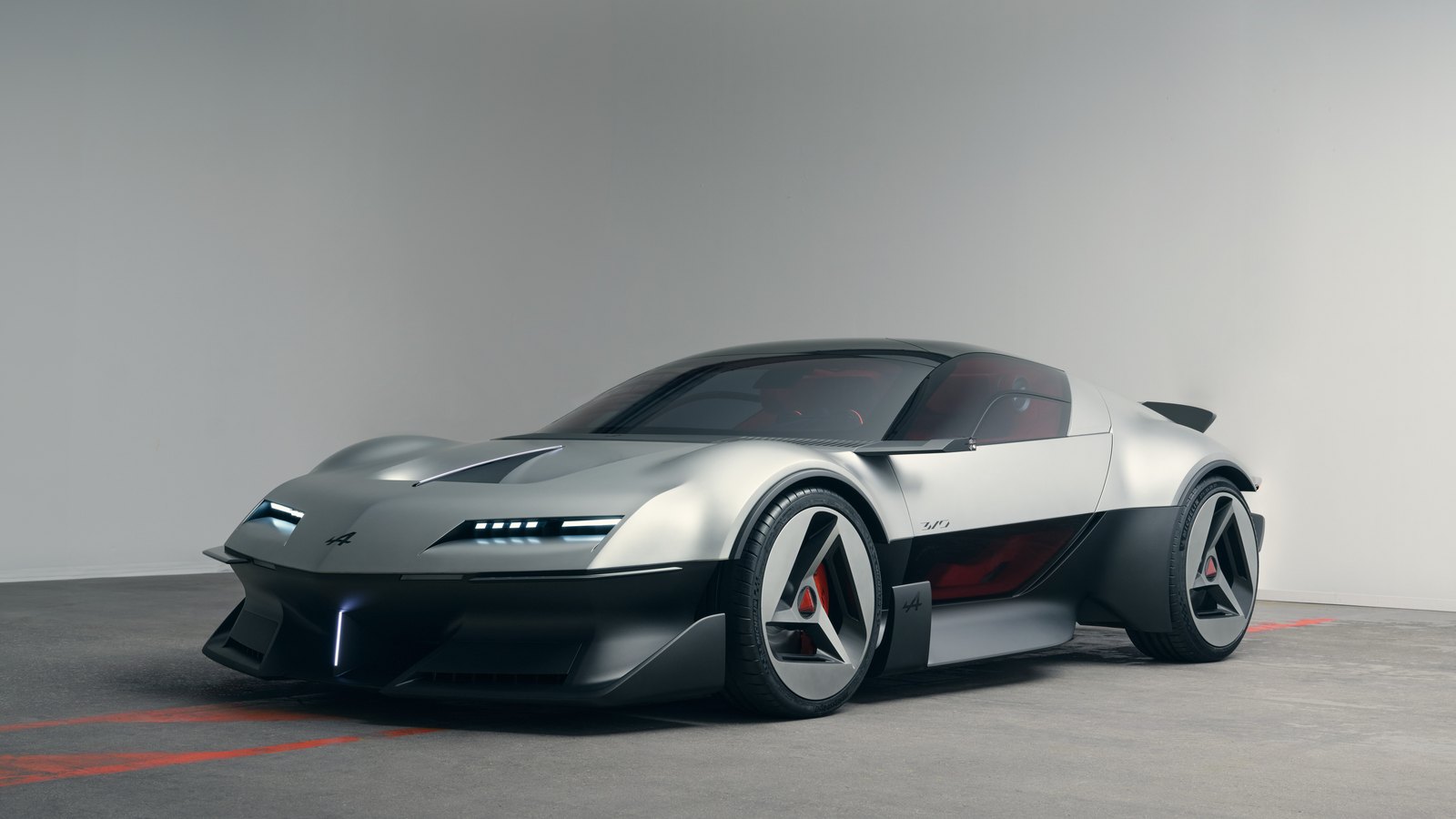 Silver and black Alpine A310 Vision concept coupe seen from the front three-quarter, with slim pop-down headlights, a sharply raked hood and triangular-spoke wheels in a gray studio