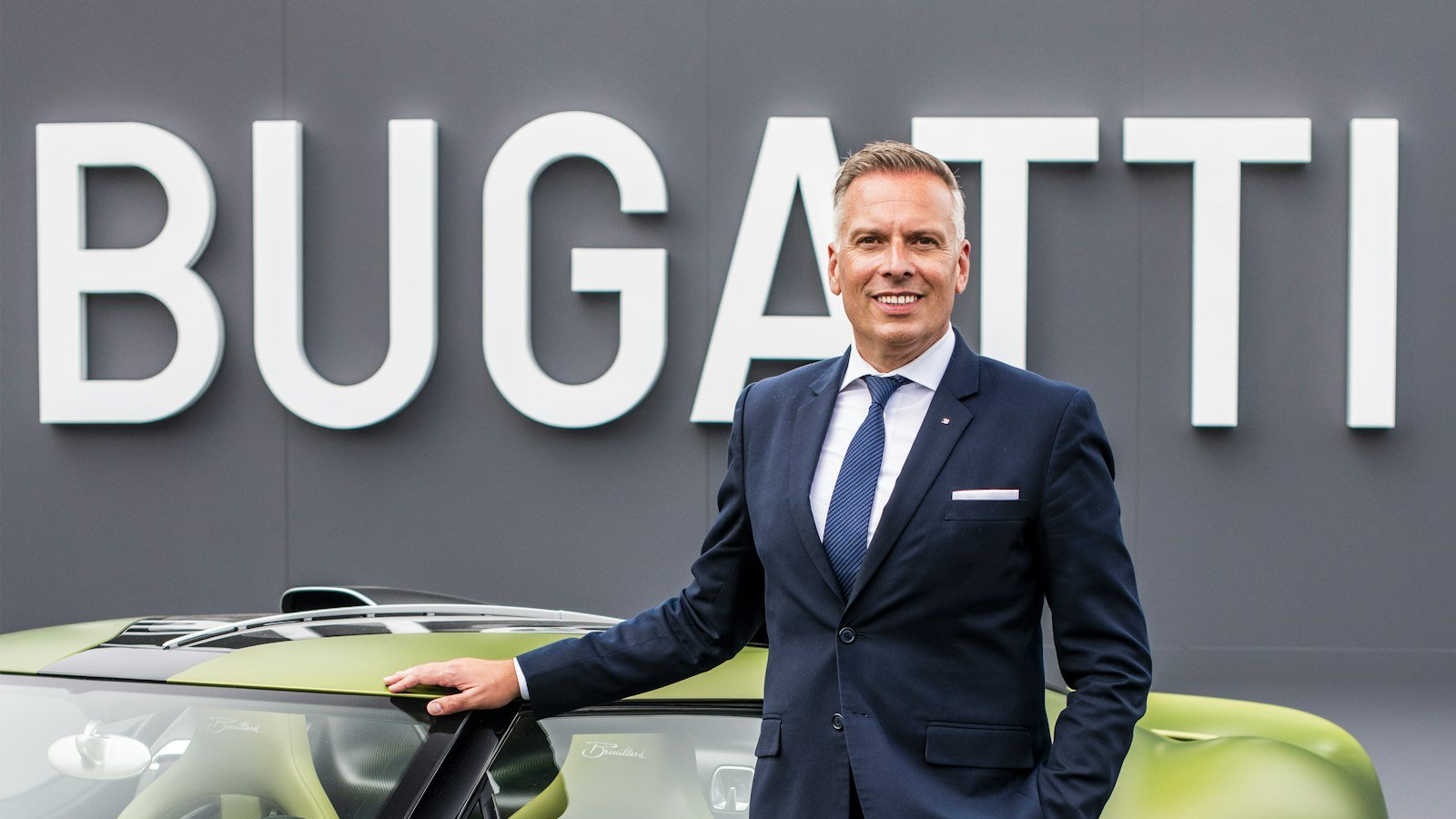 Hendrik Malinowski in a navy suit standing beside a green Bugatti in front of a large BUGATTI sign