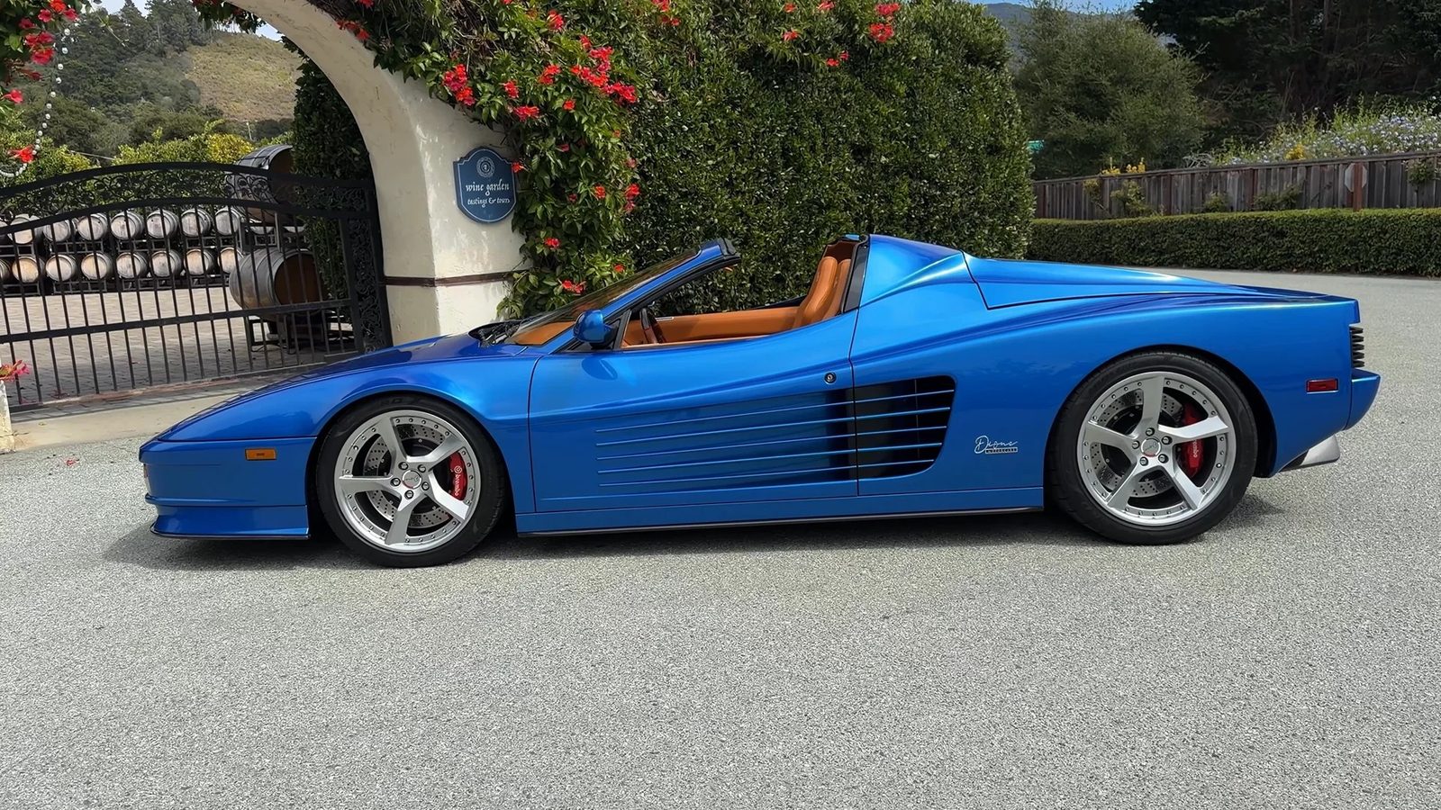 Blue Dione Tesoro in side profile with the top off, parked in front of a gated driveway