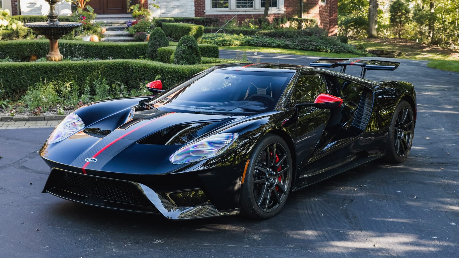 The 738-mile 2020 Ford GT Carbon Series in Shadow Black with a red center stripe, red mirror caps and black carbon-fiber wheels, parked on a driveway beside a formal hedge garden