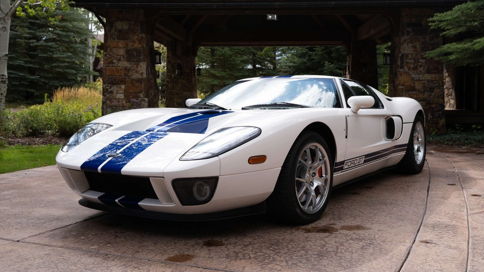 The 2k-mile 2005 Ford GT in Centennial White with blue over-the-top stripes and silver BBS wheels, parked on a stone driveway in front of a timber-and-stone carport
