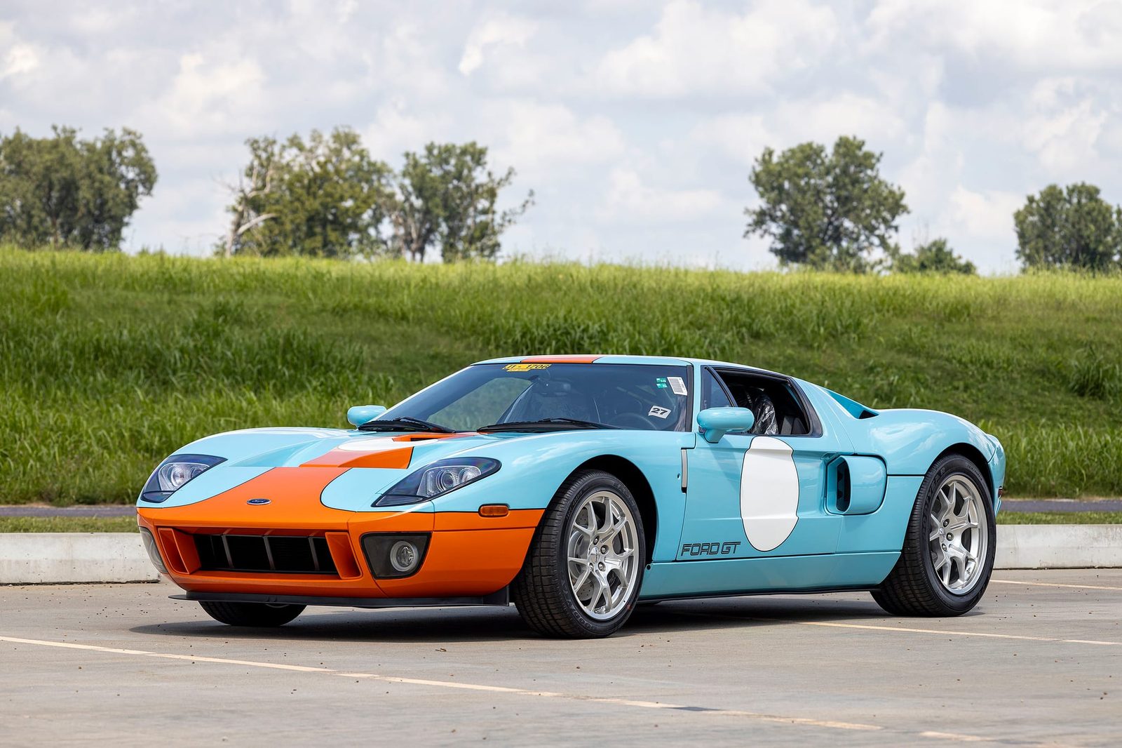 The 8-mile 2006 Ford GT Heritage Edition in light blue and orange Gulf-style colors, parked on pavement in front of a grassy bank and trees