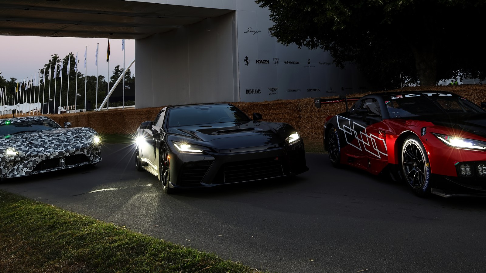 At dusk under the Goodwood Festival of Speed bridge, a camouflaged Lexus LFA prototype, a black Toyota GR GT and a red and white Toyota GR GT3 race car sit side by side with headlights on