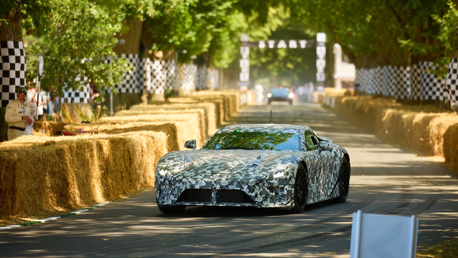 Camouflaged Lexus LFA prototype driving toward the camera on the tree-lined Goodwood hill climb, with hay bales on both sides and the START gantry behind it