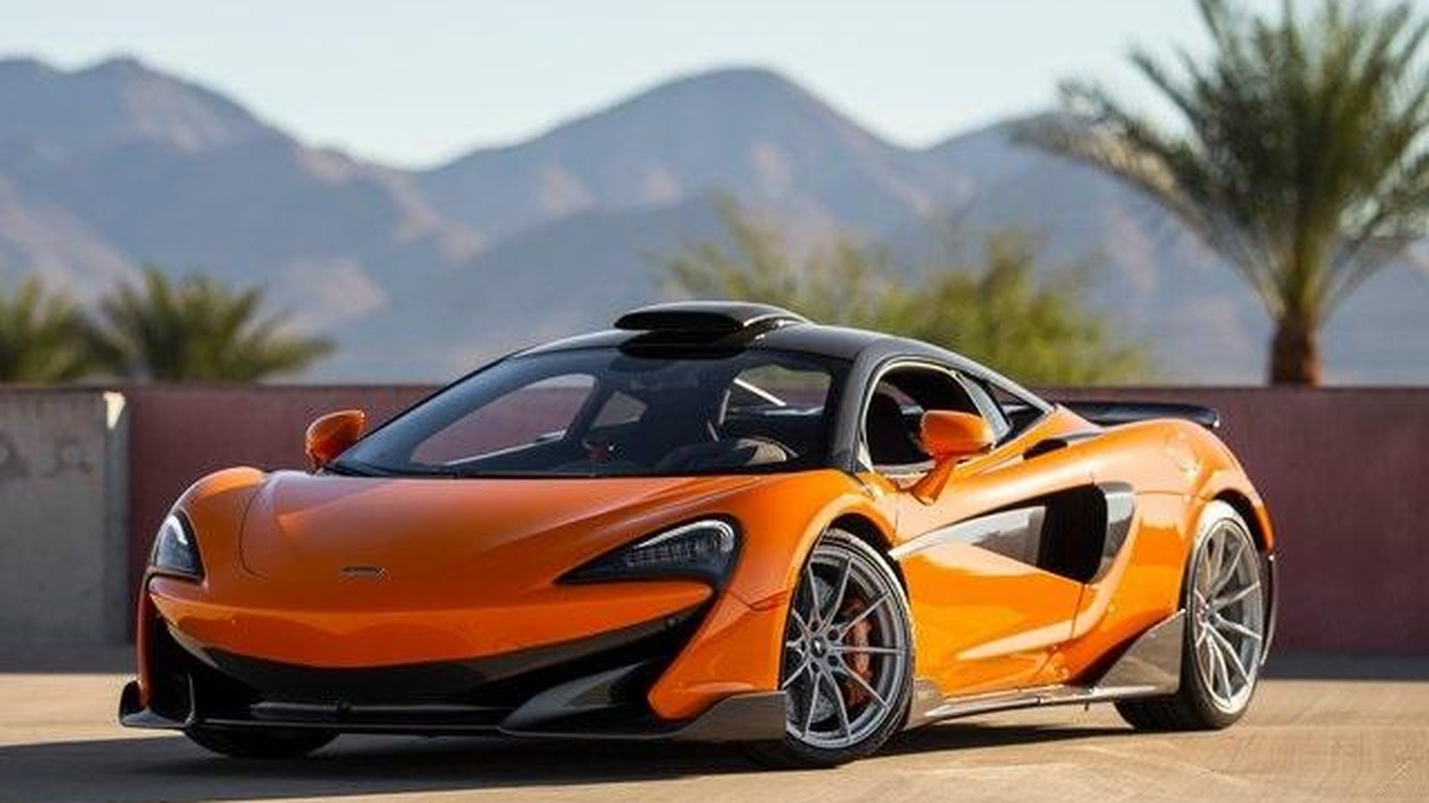 Orange McLaren 600LT coupe from the front three-quarter with mountains and palm trees behind it, the roof scoop visible above the cabin
