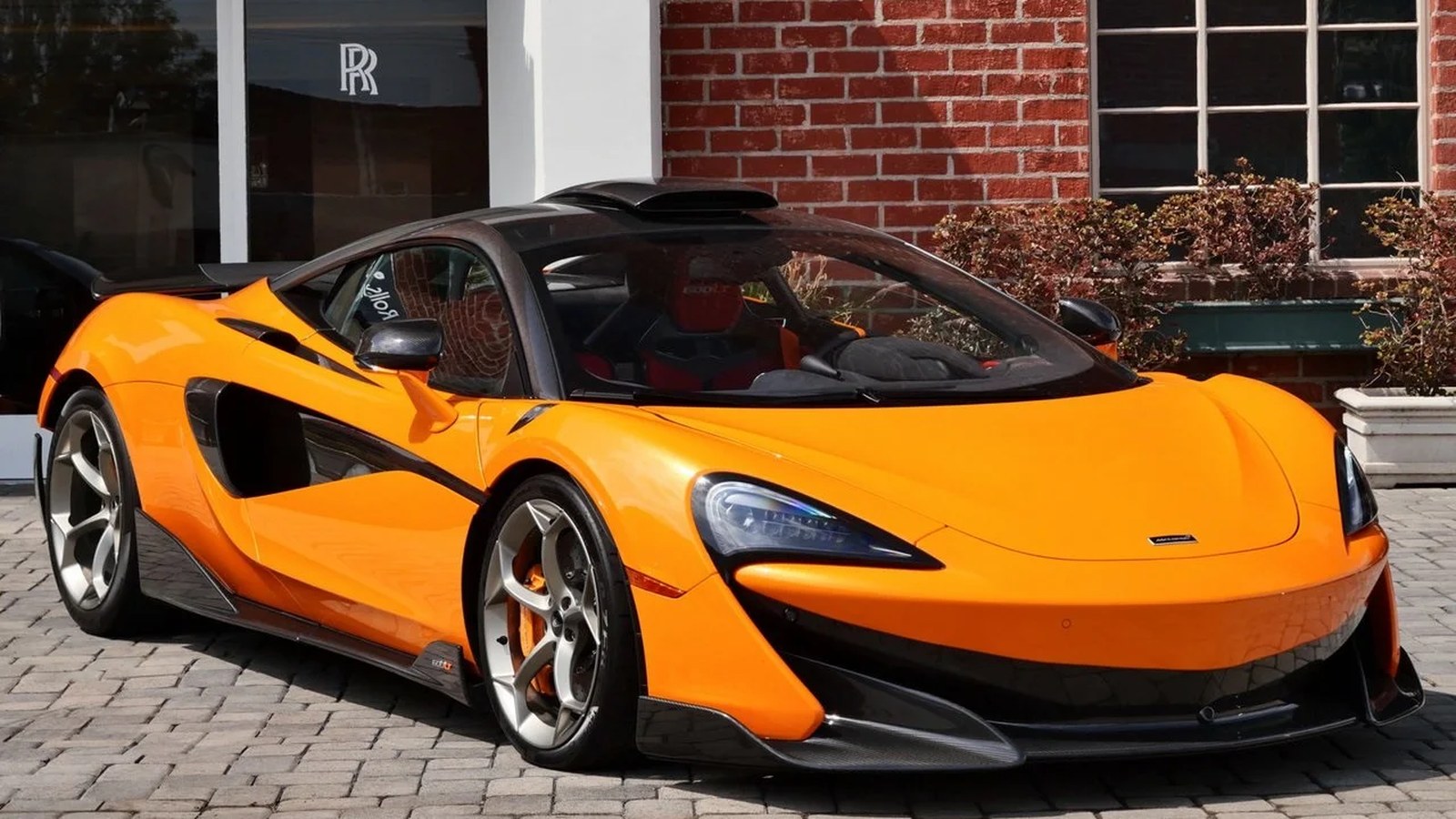 Orange McLaren 600LT coupe from the front three-quarter, parked on cobblestones outside a brick building, with the black roof scoop rising above the roof behind the windshield