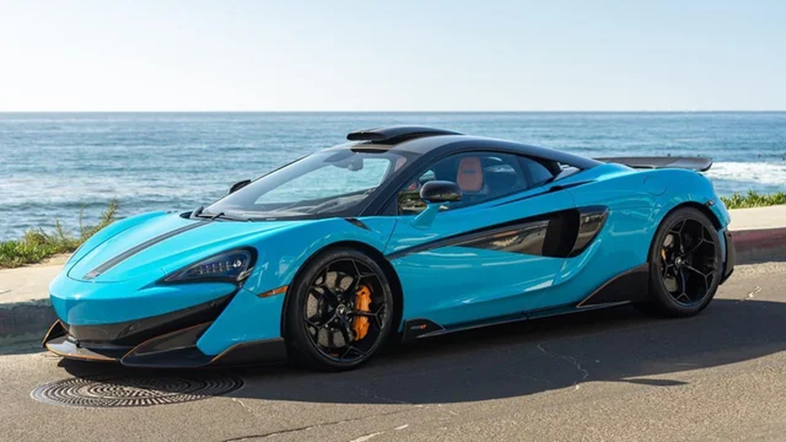 Light-blue McLaren 600LT coupe in side profile on a road above the ocean, with the roof scoop rising from the top of the roofline