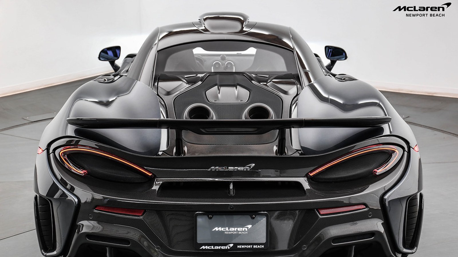 Dark grey McLaren 600LT seen straight from behind on a showroom turntable, with the roof scoop on top and the two top-exit exhaust pipes on the rear deck under the wing