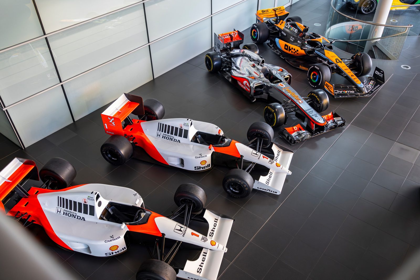 Four of the McLaren Formula 1 cars for sale lined up on a dark showroom floor: two red-and-white 1991 McLaren-Honda MP4/6As in front, with the silver 2012 MP4/27A and the papaya-and-black 2023 MCL37A behind