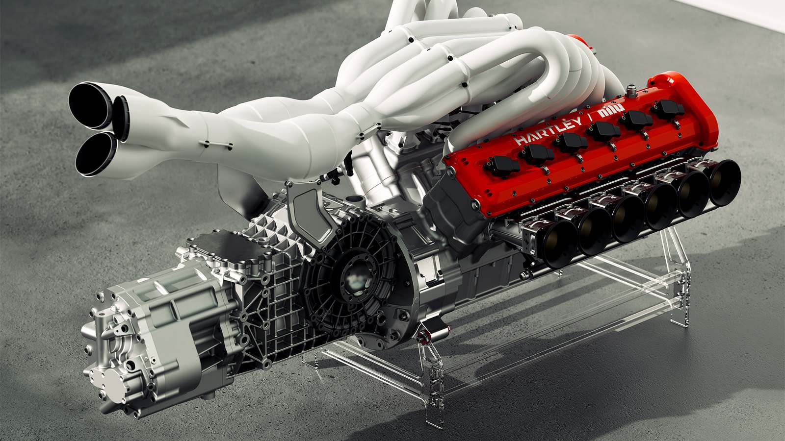 Nilu27 press image of the NILU's Hartley V12 on a clear stand, with a red cam cover reading Hartley and Nilu, a row of individual intake trumpets, white exhaust headers rising from the center and the gearbox attached at the rear