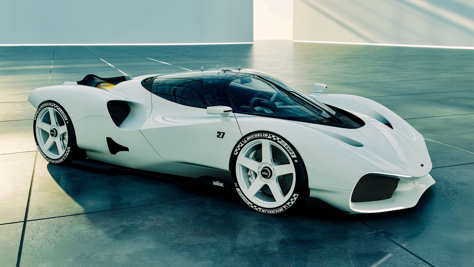 White Nilu27 NILU hypercar seen from the front three-quarter in a bright concrete hall, showing its long tapered nose, wraparound canopy, five-spoke center-lock wheels and number 27 on the door