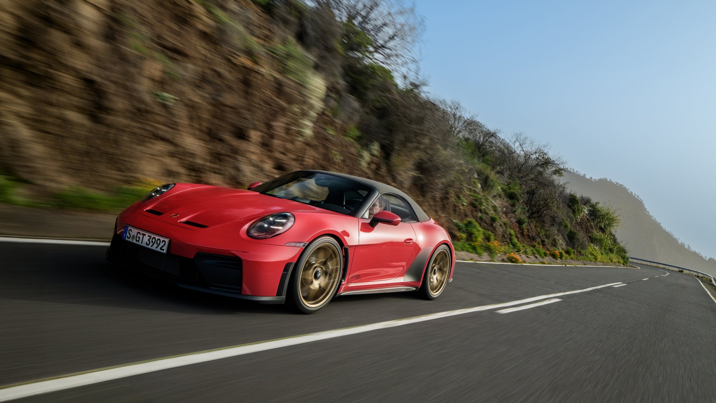 A red Porsche 911 GT3 S/C with its fabric roof up and gold wheels, driving along a mountain road, seen from the low front three-quarter angle