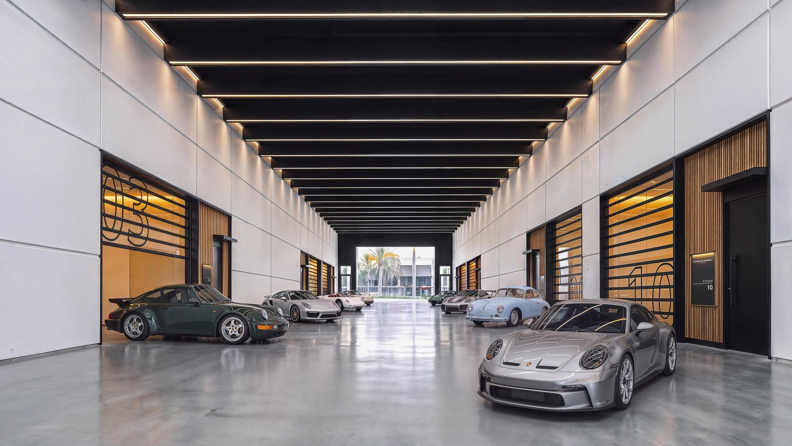 Rendering of The Concours, a long indoor driveway with a polished concrete floor and a slatted ceiling with light strips, lined with wood-and-glass studio doors, with a silver Porsche 911 in the foreground and more Porsches parked further along