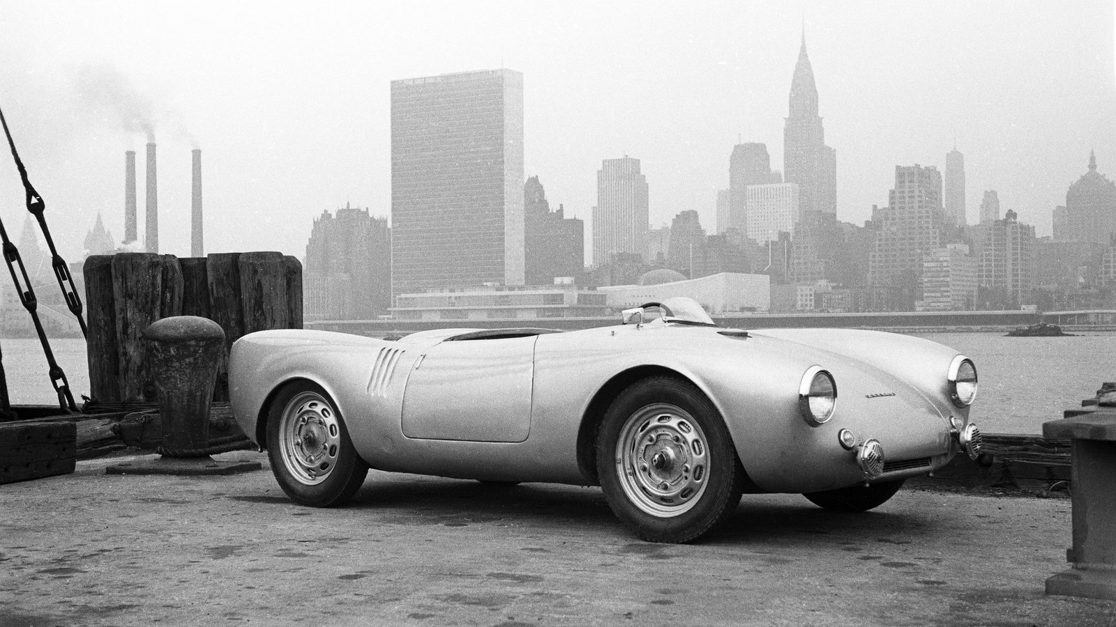 Black-and-white 1953 photo of a Porsche 550 Spyder parked on a New York pier, with the Manhattan skyline and the Chrysler Building in the background