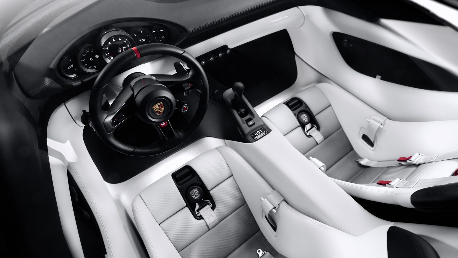 Overhead view of the Mission S cabin in white and black, showing a black steering wheel with a red top marker, five round dials behind it, no center screen, a tall gear lever on the console and two white seats with the belt buckles built into the center of each seat
